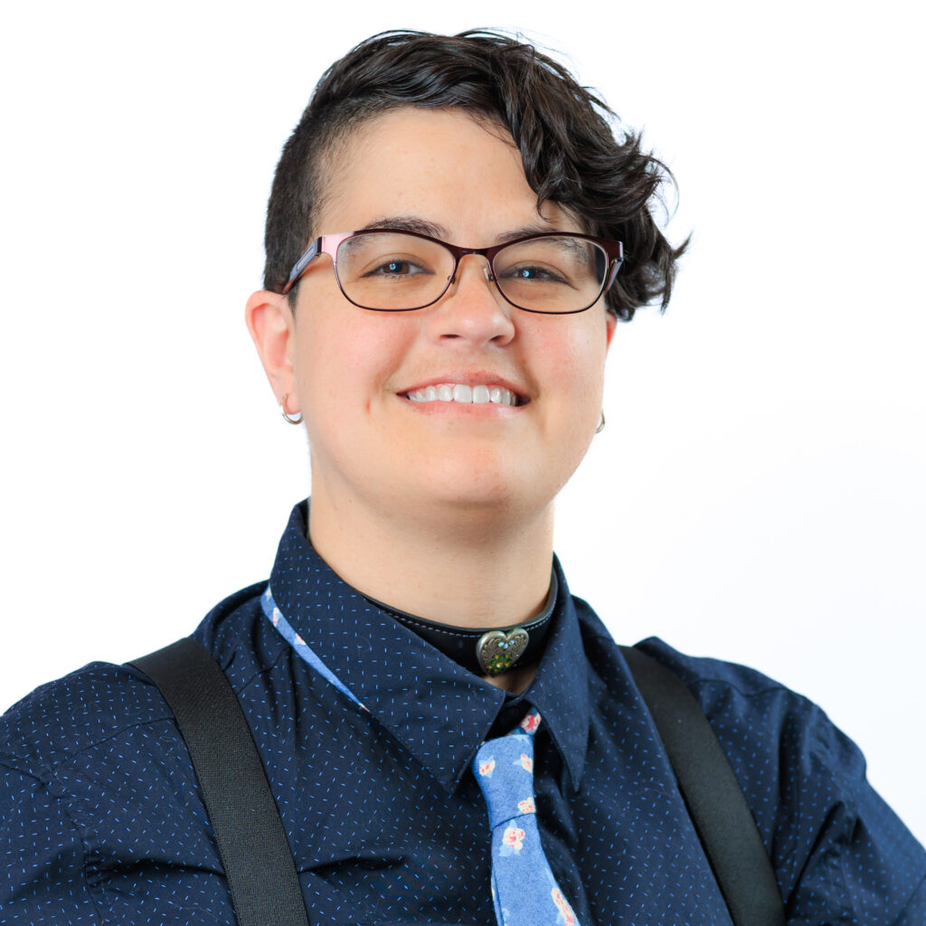 a photograph of a smiling non-binary person with short hair who is wearing glasses